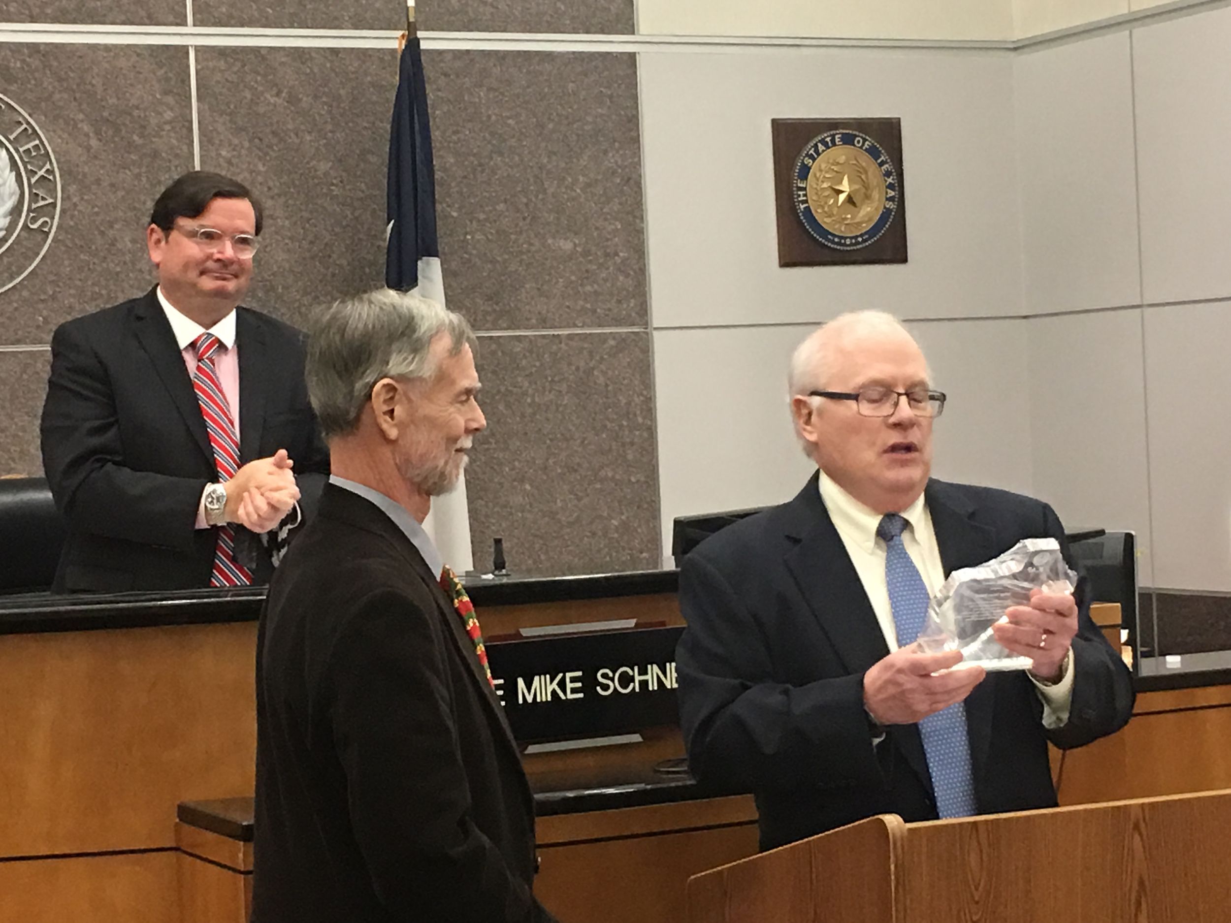Two men stand at a podium at the front of a courtroom, while a third stands behind them.