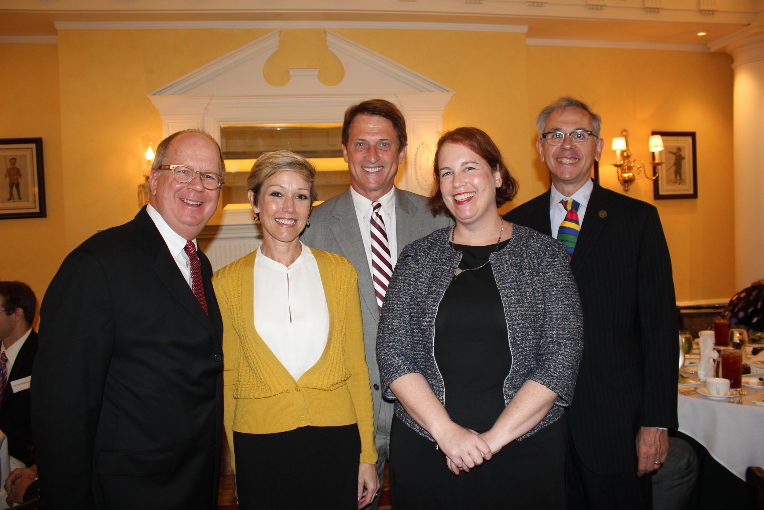 Five professionally-dressed adults stand in a group, smiling at the camera, in front of a yellow-painted wall.