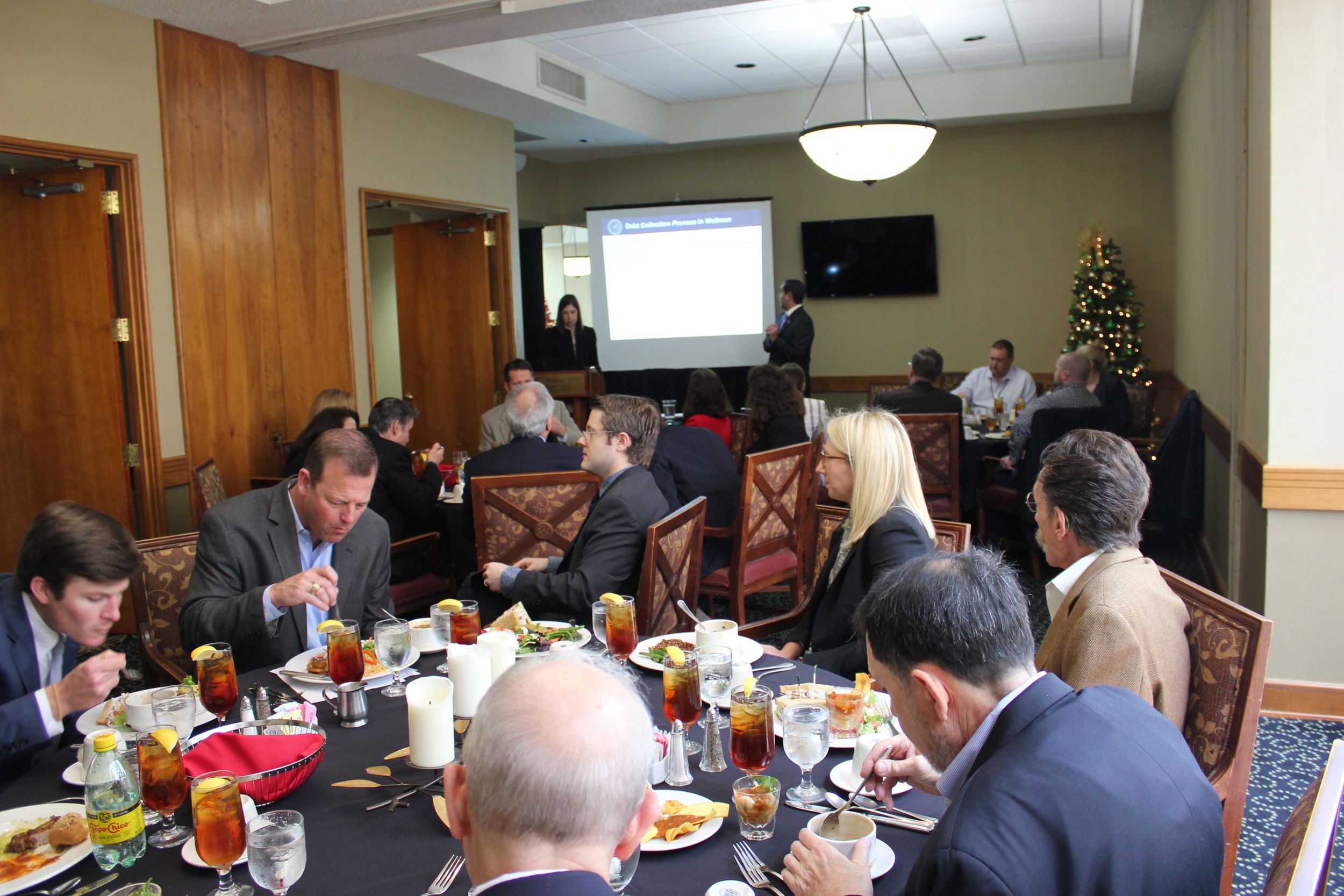 A conference room full of people eating at round tables watch a man and a woman standing in the background near a projected screen.